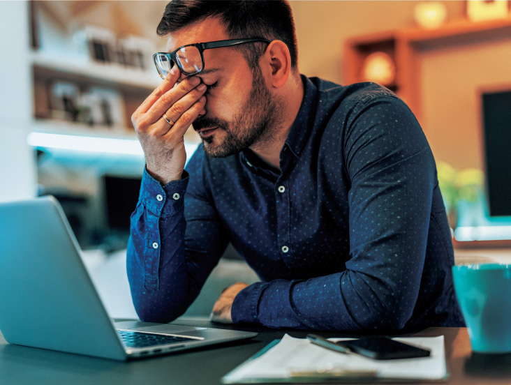 A man sitting at a desk with a laptop, holding his forehead in apparent frustration. He wears a blue shirt and glasses, and a cup and papers are on the desk. The background has warm lighting and blurred shelves.