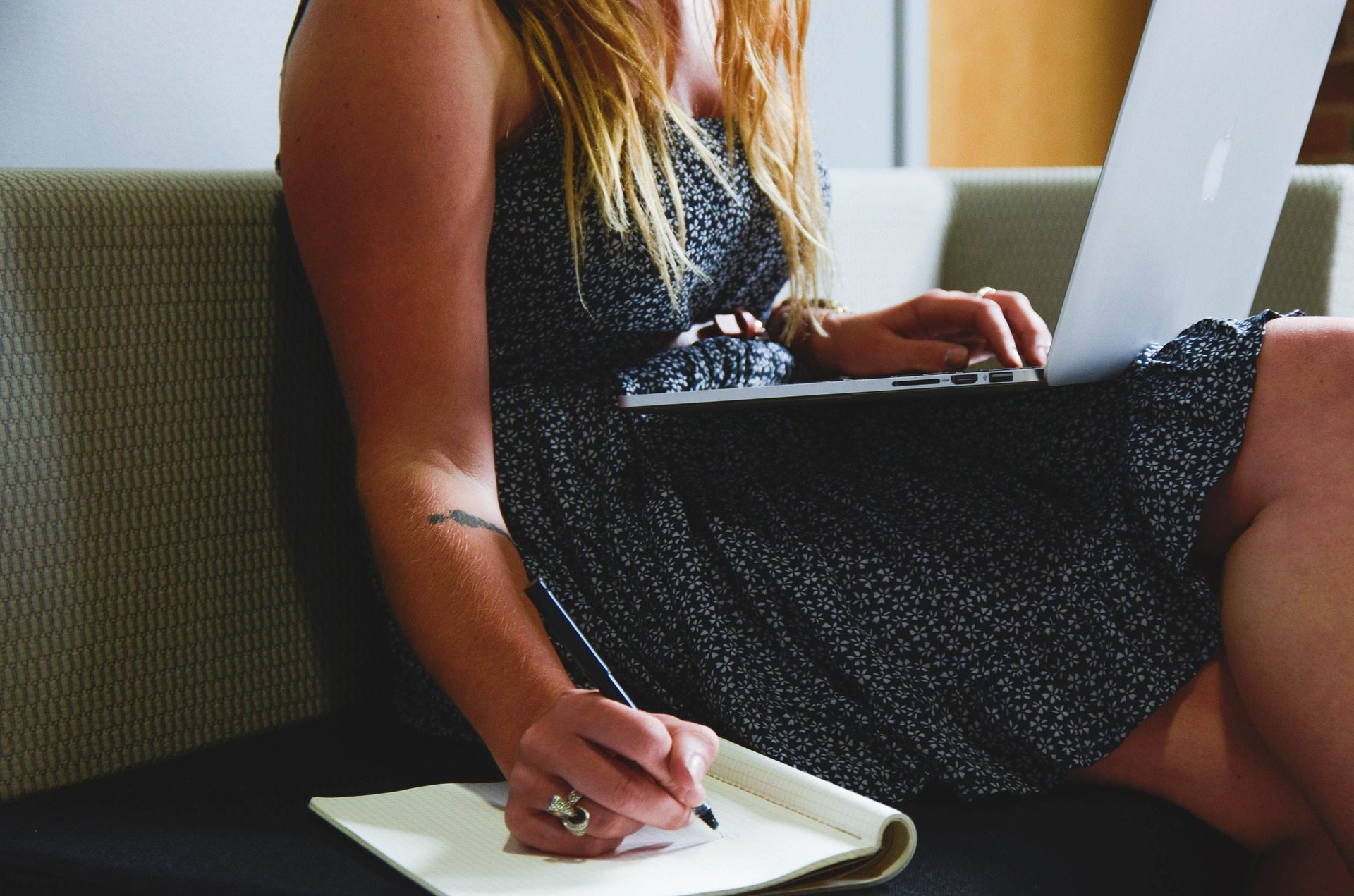 Woman sitting on couch with device on her lap, writing in a notebook next to her