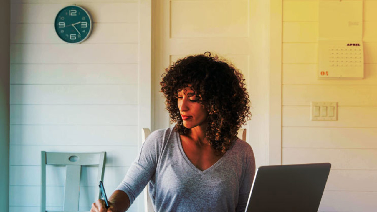 A woman sits at her dining room table with laptop and financial reports doing her monthly budget. She is writing down budgets as she works on her computer to do monthly finances, pay taxes and save money for the future.