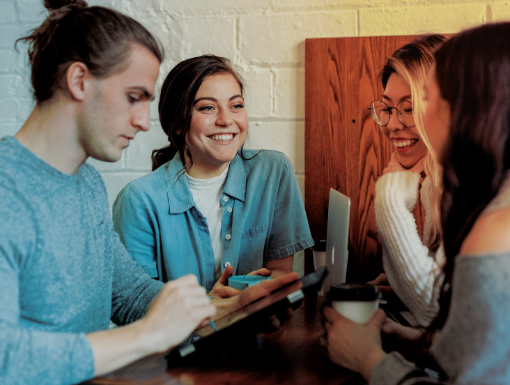 A group of four young adults sitting around a table in a cozy cafe, engaged in conversation. They are smiling and using a tablet and laptops, with one person holding a coffee cup. The background features a white brick wall.