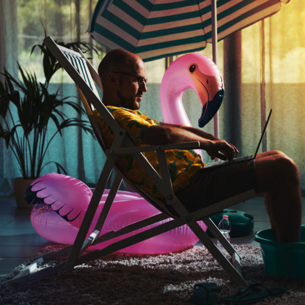 Man spending summer vacations at home alone, he is sitting on the deckchair in the living room and working with a laptop.