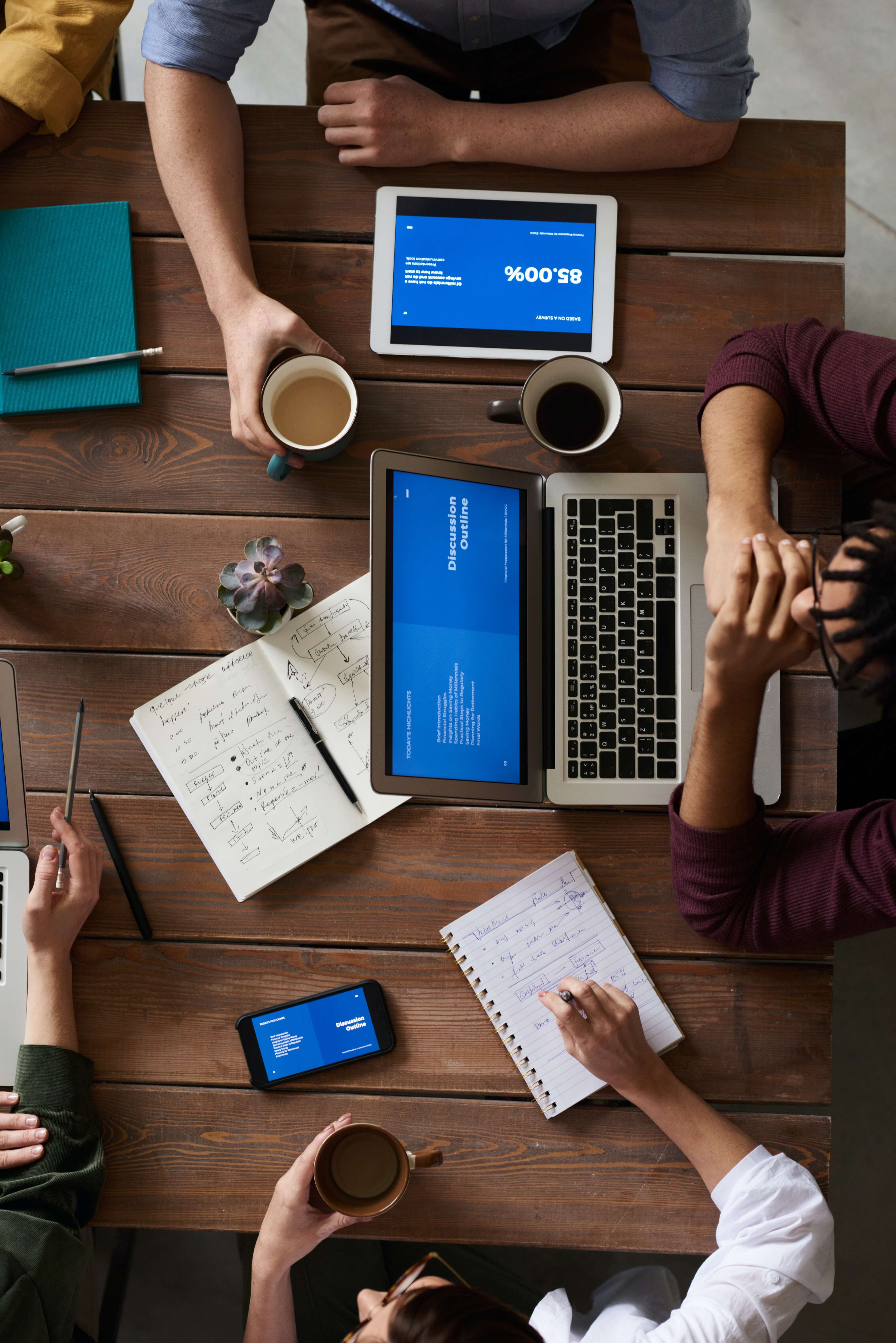 Top view of people working at a wooden desk, with their devices and notebooks