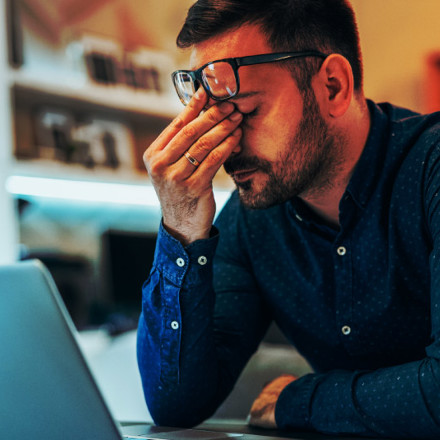 A man in a blue shirt sits at a desk with his laptop open. He leans forward, resting his elbow on the desk, and pinches the bridge of his nose with one hand, appearing stressed or fatigued.