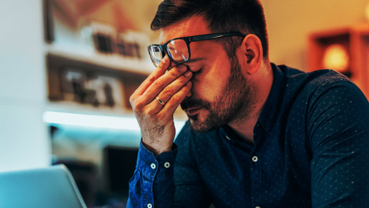 A man in a blue shirt sits at a desk with his laptop open. He leans forward, resting his elbow on the desk, and pinches the bridge of his nose with one hand, appearing stressed or fatigued.