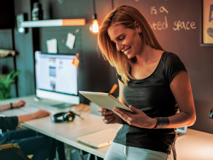 A woman in a black t-shirt is smiling while using a tablet. She is leaning against a desk in a modern office with a computer monitor in the background. The setting has warm lighting and creative decor.