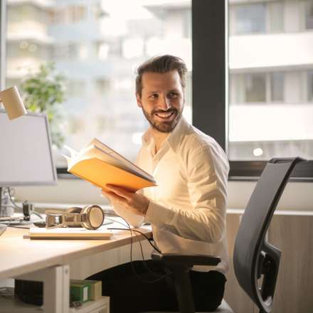 Smiling man holding yellow book, turns around in desk seat and looks back